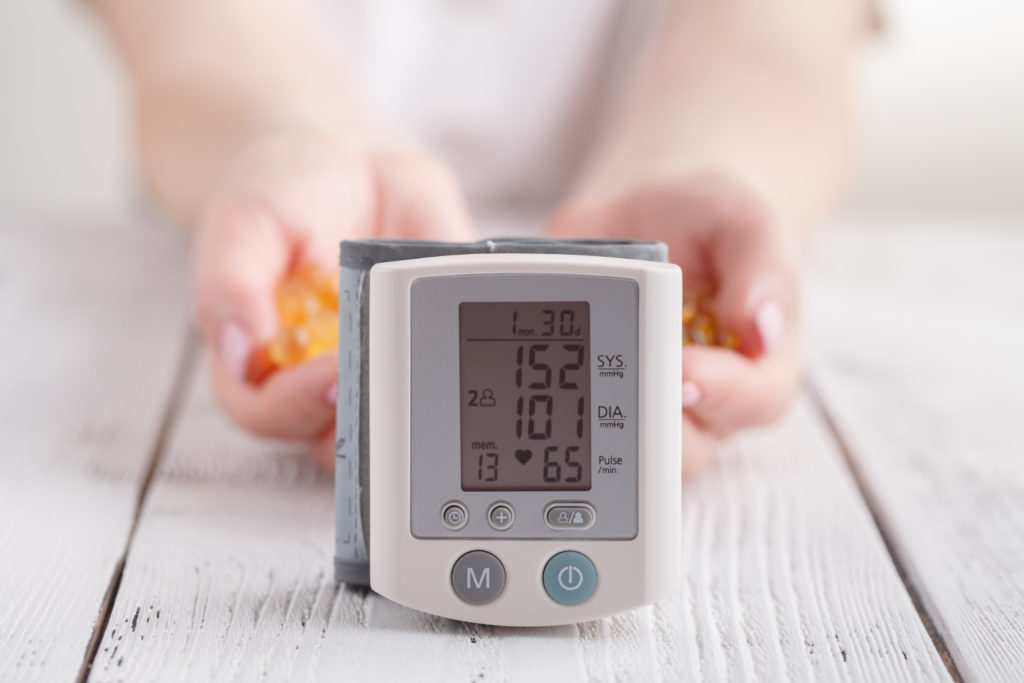 close up of blood pressure monitor with hands holding capsules of fish oil and vitamin e